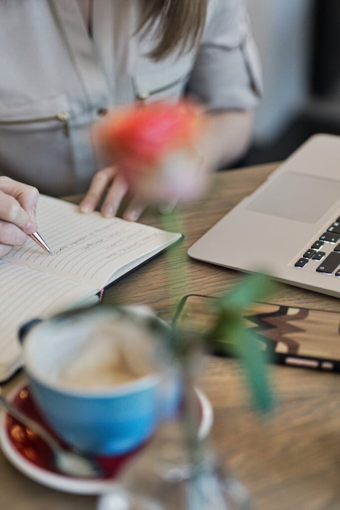Woman writing in a notebook with a laptop and coffee cup on a desk. Ideal for workspace inspiration.